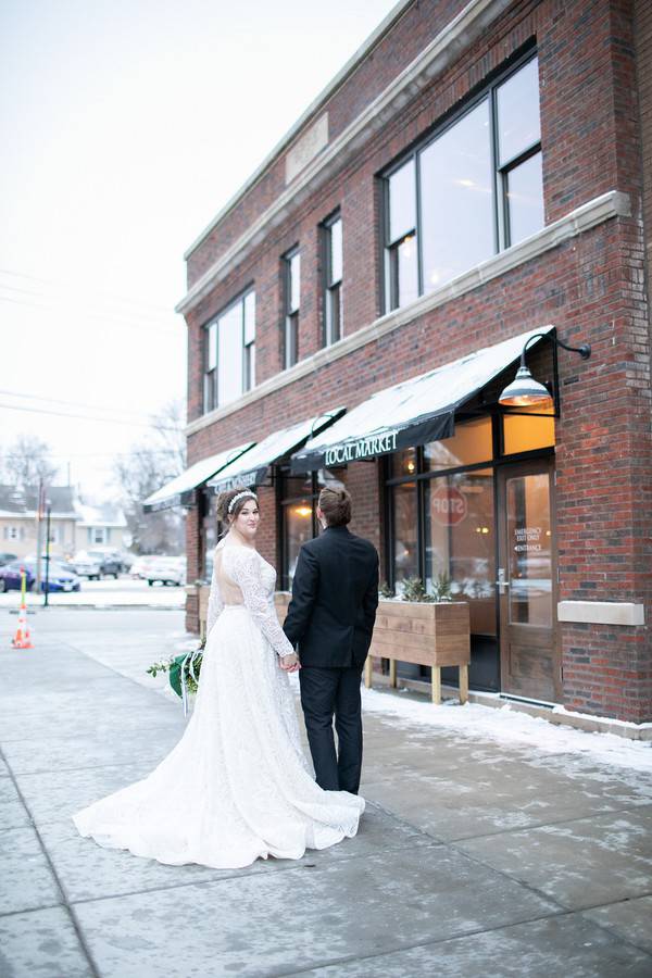 Bride in ivory lace A-line wedding dress with long lace sleeves and open back walking hand-in-hand with groom on a snowy urban street