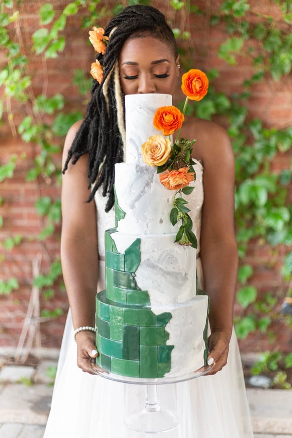 Bride holding a four-tier wedding cake with hand-painted green botanical artwork, tropical leaves, and peach flowers on a textured ivory surface