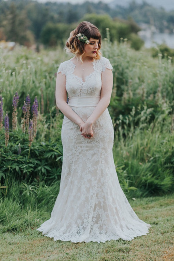 Bride Caryn in an ivory cap-sleeve lace mermaid gown with V-neckline and ribbon sash, standing in a wildflower garden at Kildara Farms in North Saanich, British Columbia