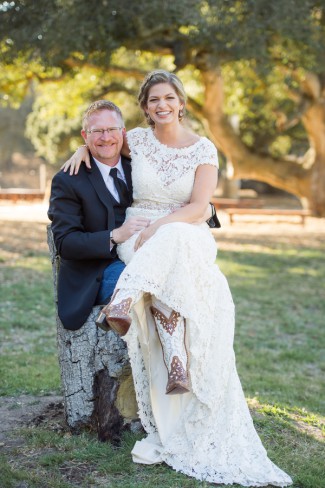 Bride in lace cap-sleeve wedding gown sitting on groom's knee, showing off cowboy boots, at California ranch wedding