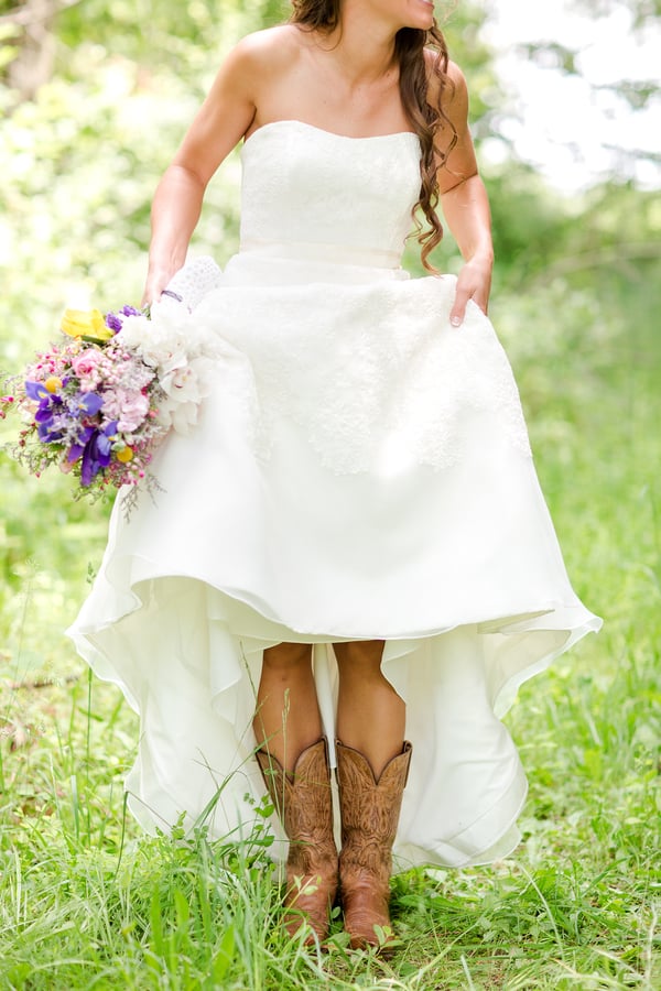 Bride in strapless a-line wedding gown lifting the skirt to show brown cowboy boots, holding colorful wildflower bouquet, outdoors in green grass