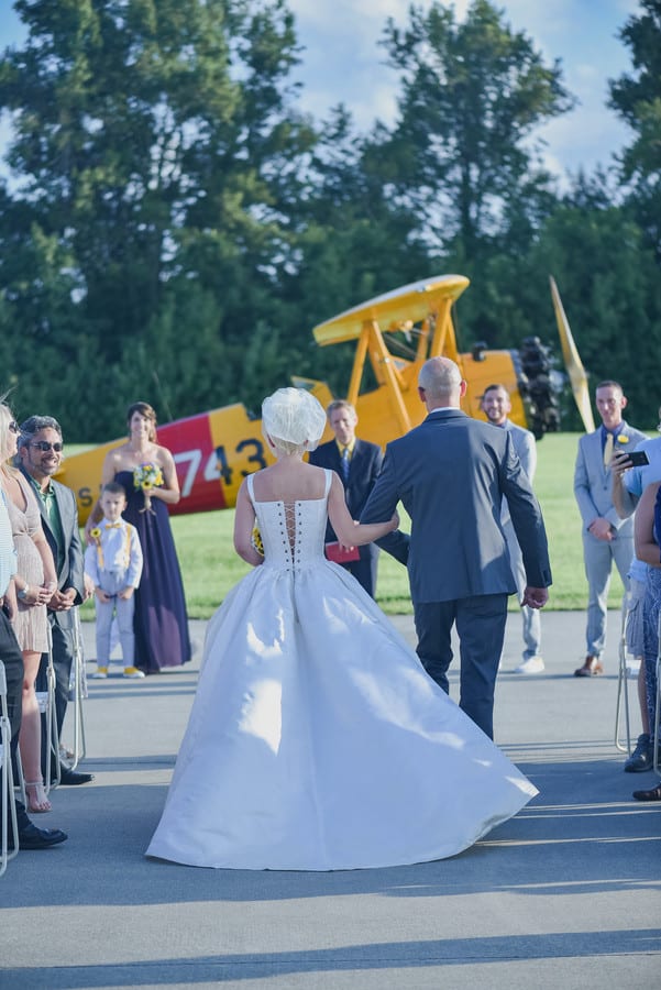 Bride Heidi in a white corset ballgown with birdcage fascinator walking up a tarmac aisle with her father at the Military Aviation Museum in Virginia Beach, Virginia, with a vintage yellow biplane in the background