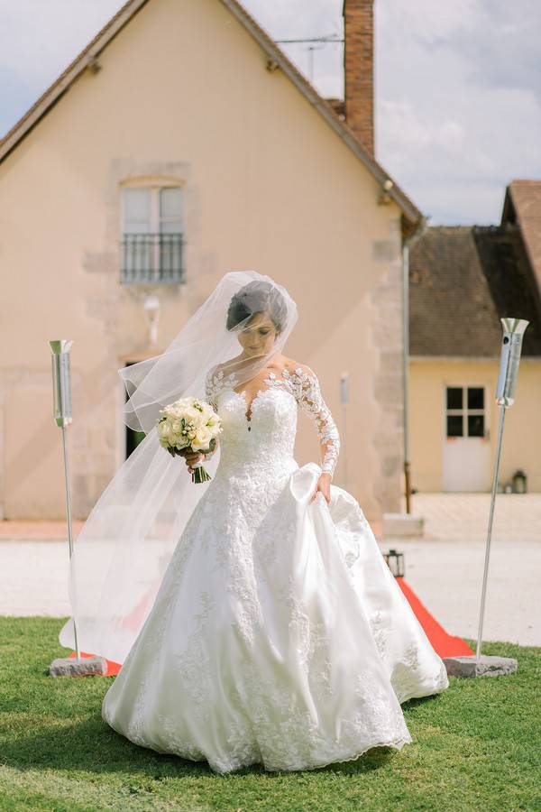 Bride walking up outdoor aisle in full lace ballgown with illusion long sleeves and massive tulle skirt at French chateau wedding