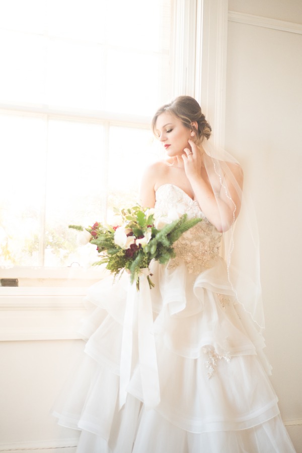 Bride in strapless sweetheart lace and tulle ruffled tiered ballgown holding lush Christmas bouquet by window at Liriodendron Mansion in Maryland
