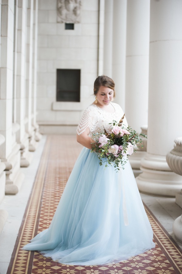 Bride in two-piece wedding outfit with white lace crop top with flutter sleeves and a powder blue tulle ball gown skirt holding a pink bouquet in a marble corridor