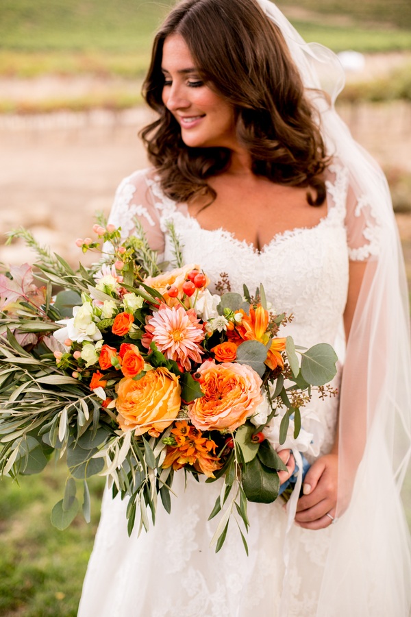Bride at a vineyard smiling down at a large organic fall bouquet of orange and peach garden roses, café au lait dahlias, and trailing olive and eucalyptus branches