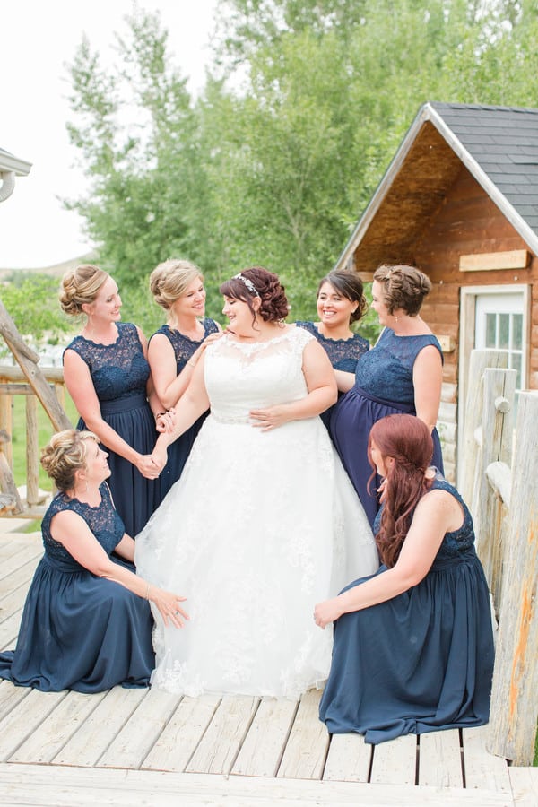 Plus-size bride in white ball gown with illusion lace neckline and lace bodice surrounded by bridesmaids in navy lace dresses on an outdoor deck