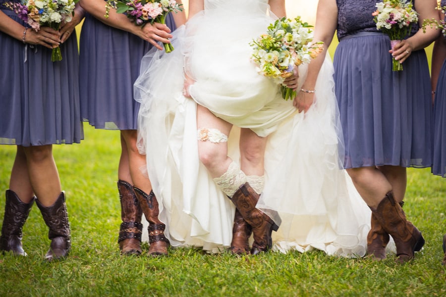 Bride in white ballgown lifting dress to show cowboy boot and lace garter, with bridesmaids in violet dresses and cowboy boots, at Carlee Farm wedding
