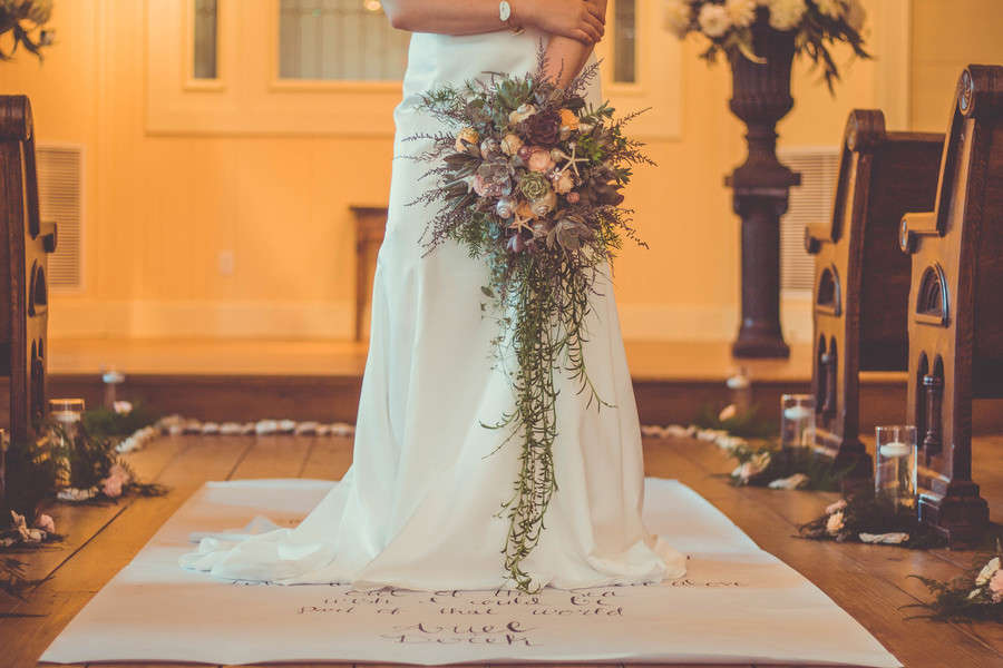 Bride standing in a candlelit church aisle holding a long cascading bouquet with blush roses at the top and trailing fern greenery reaching nearly to her gown hem