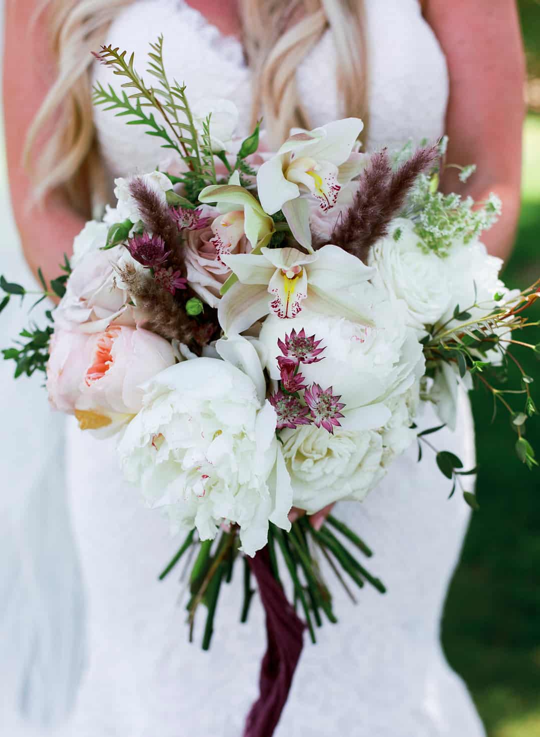 Close-up of a lush bridal bouquet featuring giant white peonies, white cymbidium orchid sprays, blush garden roses, and star-shaped astrantia with a deep burgundy ribbon wrapping the stems