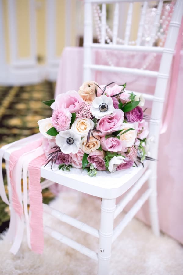 Lush pink and blush bridal bouquet with layered garden roses, white anemones with black centers, and clusters of ranunculus resting on a white Chiavari chair draped in blush ribbon