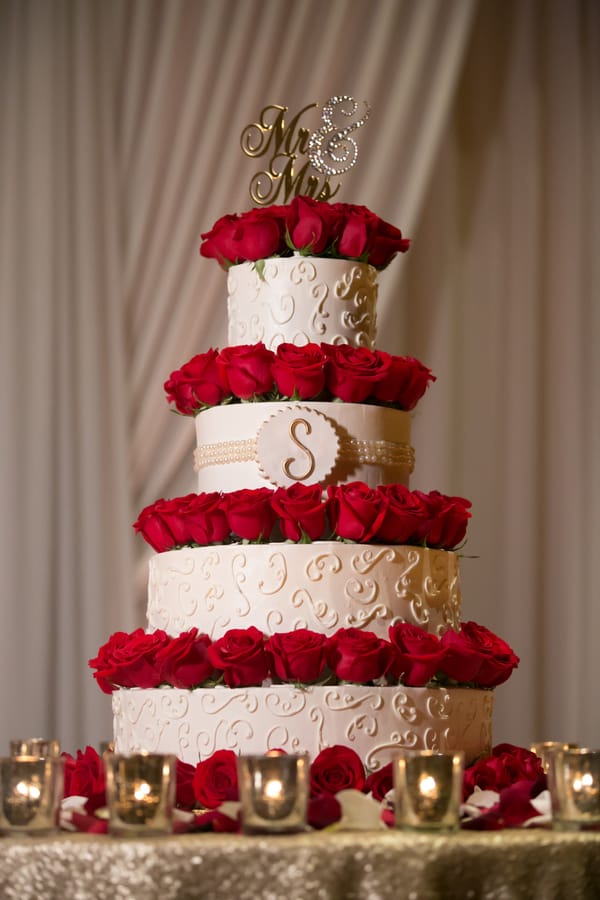Four-tier cream wedding cake with rings of deep red roses around each tier, a gold initial monogram on the front, and a cake topper