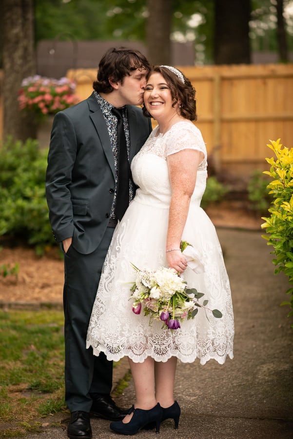 Plus-size bride in white tea-length lace dress with cap sleeves and full A-line skirt in a backyard garden setting with groom