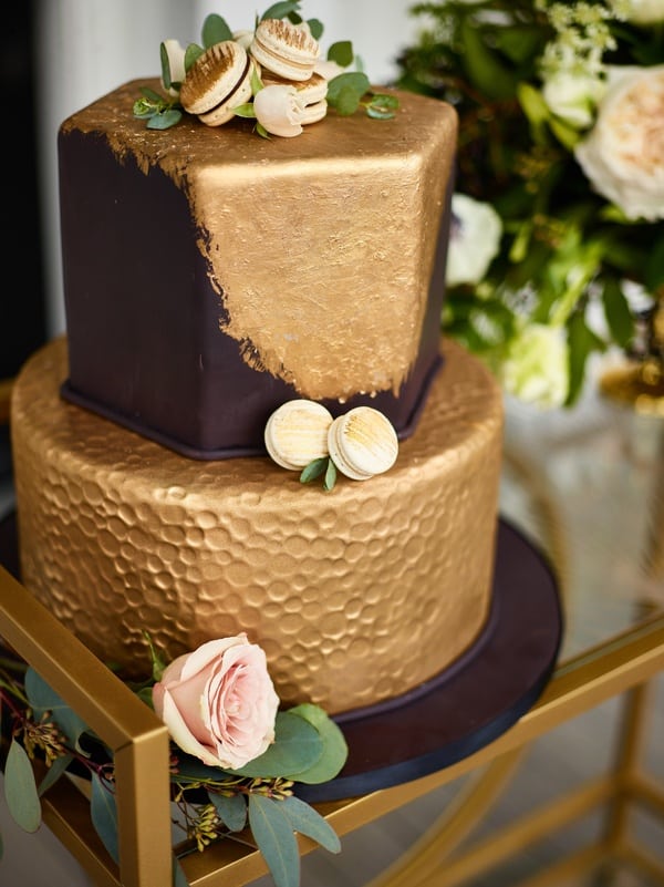 Two-tier hexagonal wedding cake with hammered gold fondant texture on the lower tier and a dark second tier with white floral accents