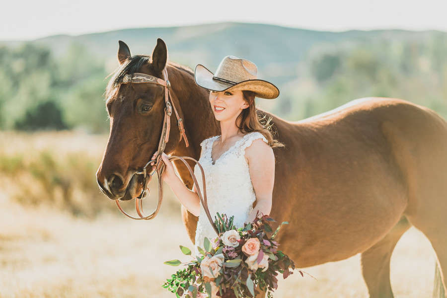 Bride in fitted lace bridal gown wearing a straw cowboy hat, standing beside a brown horse with colorful bouquet in a golden California field