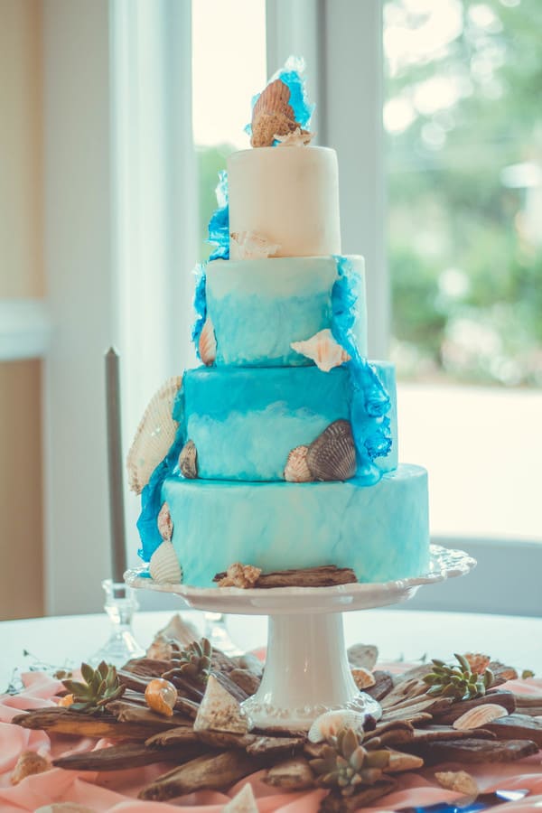 Four-tier blue fondant wedding cake with hand-painted underwater sea life scenes, ocean waves, and a seascape in teal and aqua