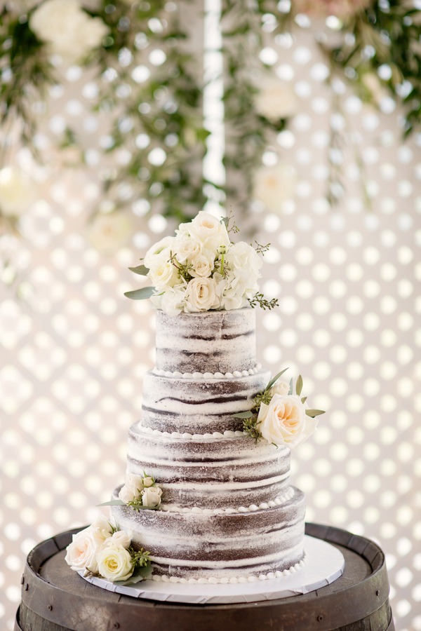 Four-tier semi-naked wedding cake draped in white garden roses, ranunculus, and eucalyptus against a lace backdrop