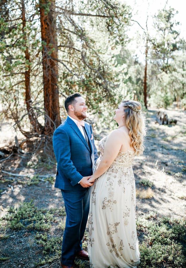 Bride in champagne gold beaded A-line gown with spaghetti straps and V-neckline standing in a sunny pine forest with groom