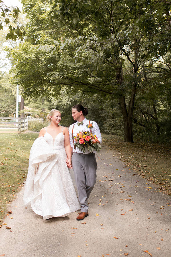 Bride in ivory lace A-line wedding dress with spaghetti straps and V-neckline walking with groom through a tree-lined park path