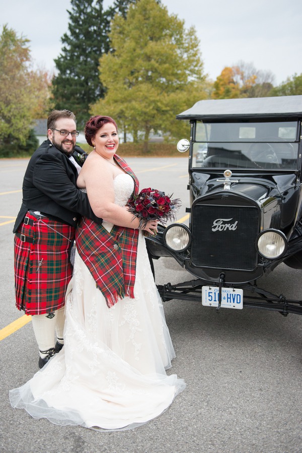 Plus-size bride in ivory strapless sweetheart ball gown with lace bodice and tulle skirt beside a vintage Ford car