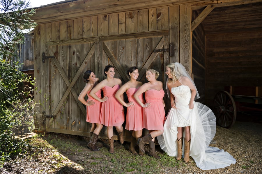 Bride in strapless white a-line wedding gown with bridesmaids in coral dresses and cowboy boots in front of rustic barn at Fair Weather Farms