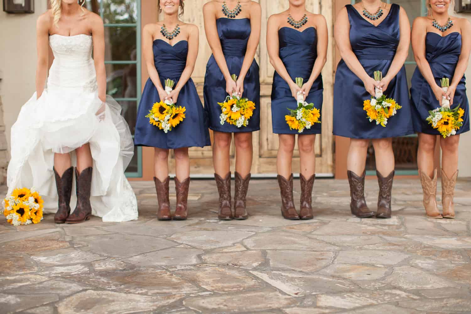 Bride in strapless white high-low wedding gown with bridesmaids in navy blue dresses and cowboy boots holding sunflower bouquets at Robinson Ranch Golf Club California
