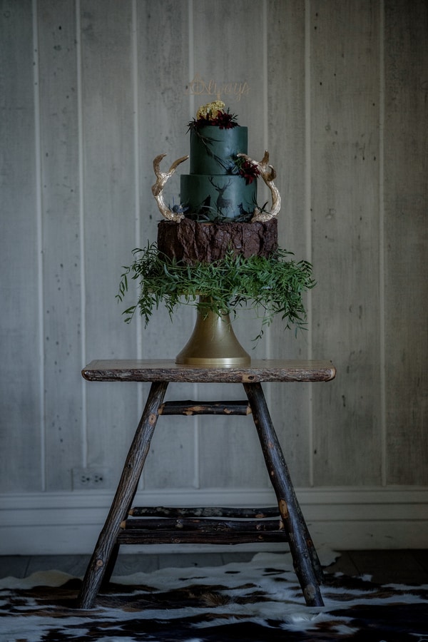 Three-tier dark forest green wedding cake with hand-painted botanical details, gold stag antlers, and cascading greenery on a gold pedestal