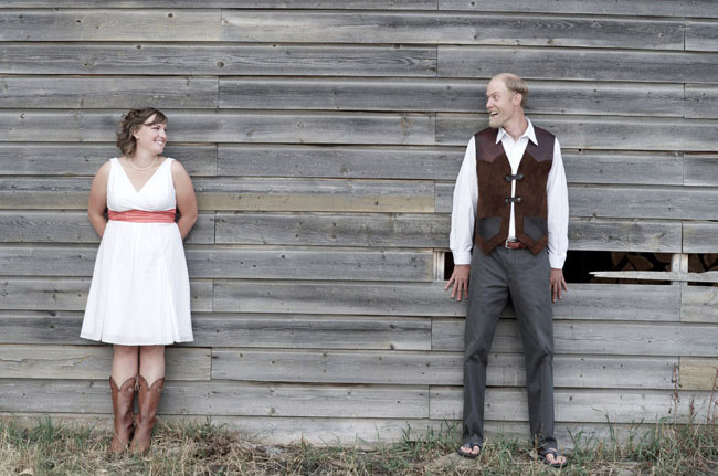 Bride in white sleeveless V-neck midi wedding dress with coral sash and cowboy boots standing beside groom against a barn wall in Montana