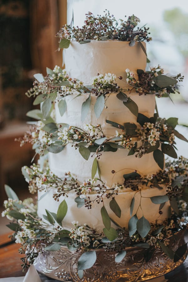 Four-tier wedding cake with textured unfinished buttercream icing and cascading eucalyptus vines, dark berries, and small white florals