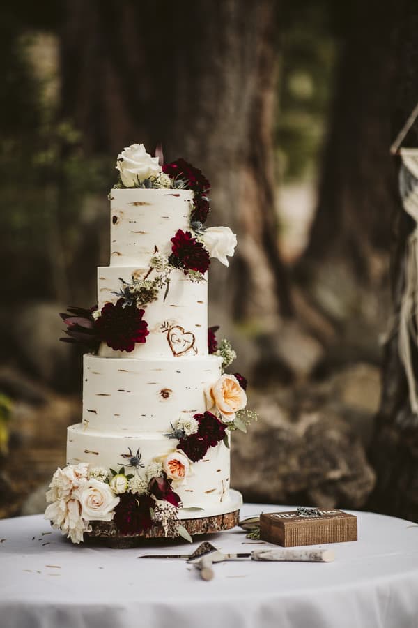 Four-tier white birch bark-textured wedding cake with a carved J+J heart, surrounded by dark burgundy roses and lush greenery