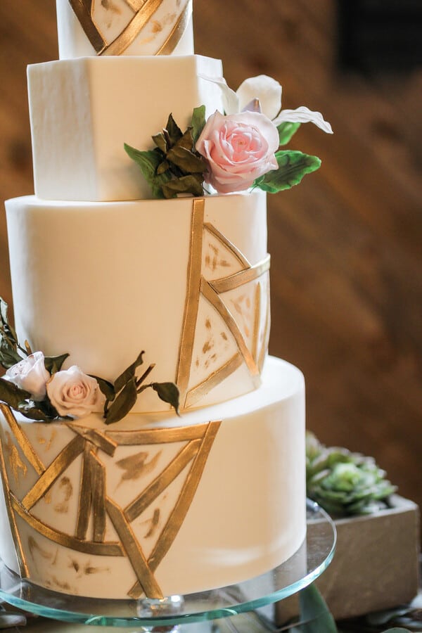 Four-tier wedding cake with mixed geometric tier shapes including round and pentagon tiers, decorated with gold architectural line work