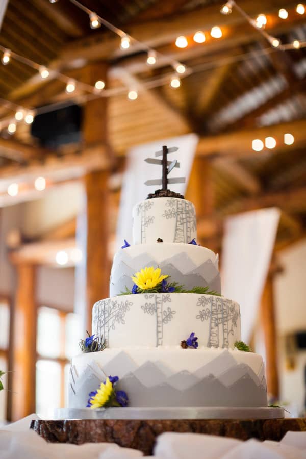Four-tier white fondant wedding cake with a miniature mountain forest landscape sculpted onto the tiers, including evergreen trees and a wooden topper