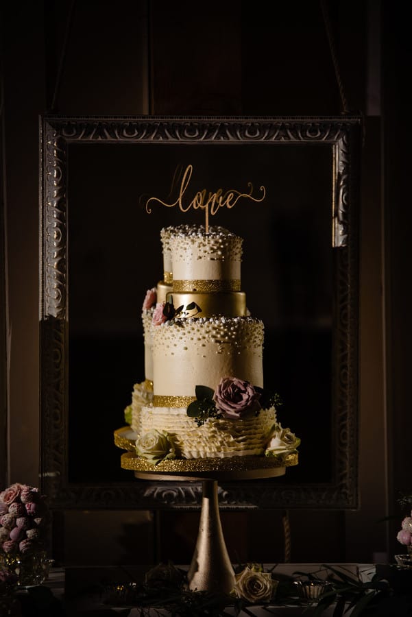 Three-tier wedding cake with a black fondant base tier covered in gold glitter confetti, ivory lace-patterned tiers, and a purple floral accent