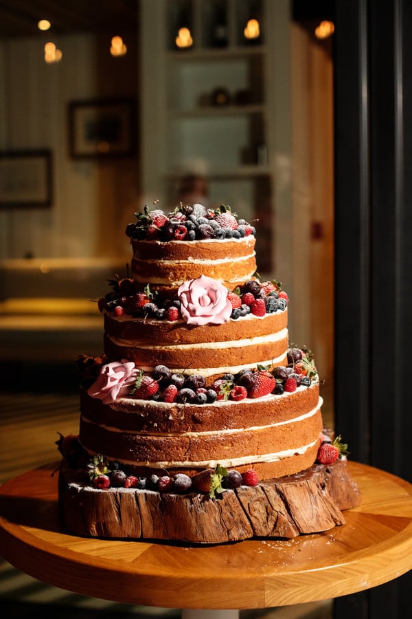 Three-tier naked wedding cake piled high with fresh red berries, dark fruit, and a blush garden rose on top