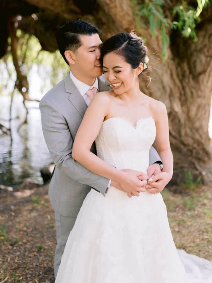 Groom standing behind the bride with arms around her waist, both facing the same way by a tree