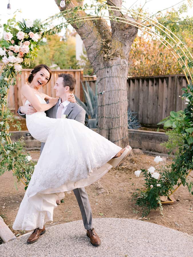 Groom lifting and carrying the laughing bride in his arms in front of a floral arch