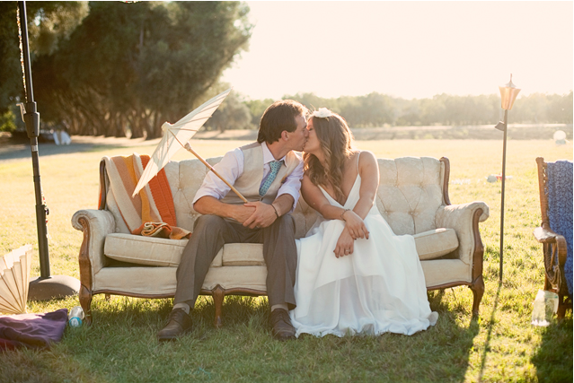 Bride and groom sitting on a vintage couch in a golden field, leaning into a kiss