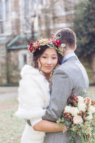 Bride in a fur wrap and berry flower crown hugging the groom, her face turned toward the camera