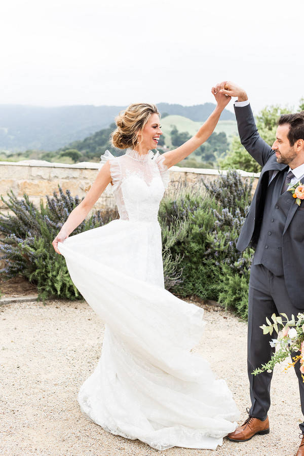 Groom spinning the bride by one raised joined hand, her lace dress flaring on a hillside