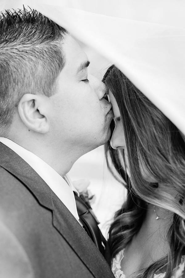 Black-and-white close-up of the groom kissing the bride's temple, her veil draping over both