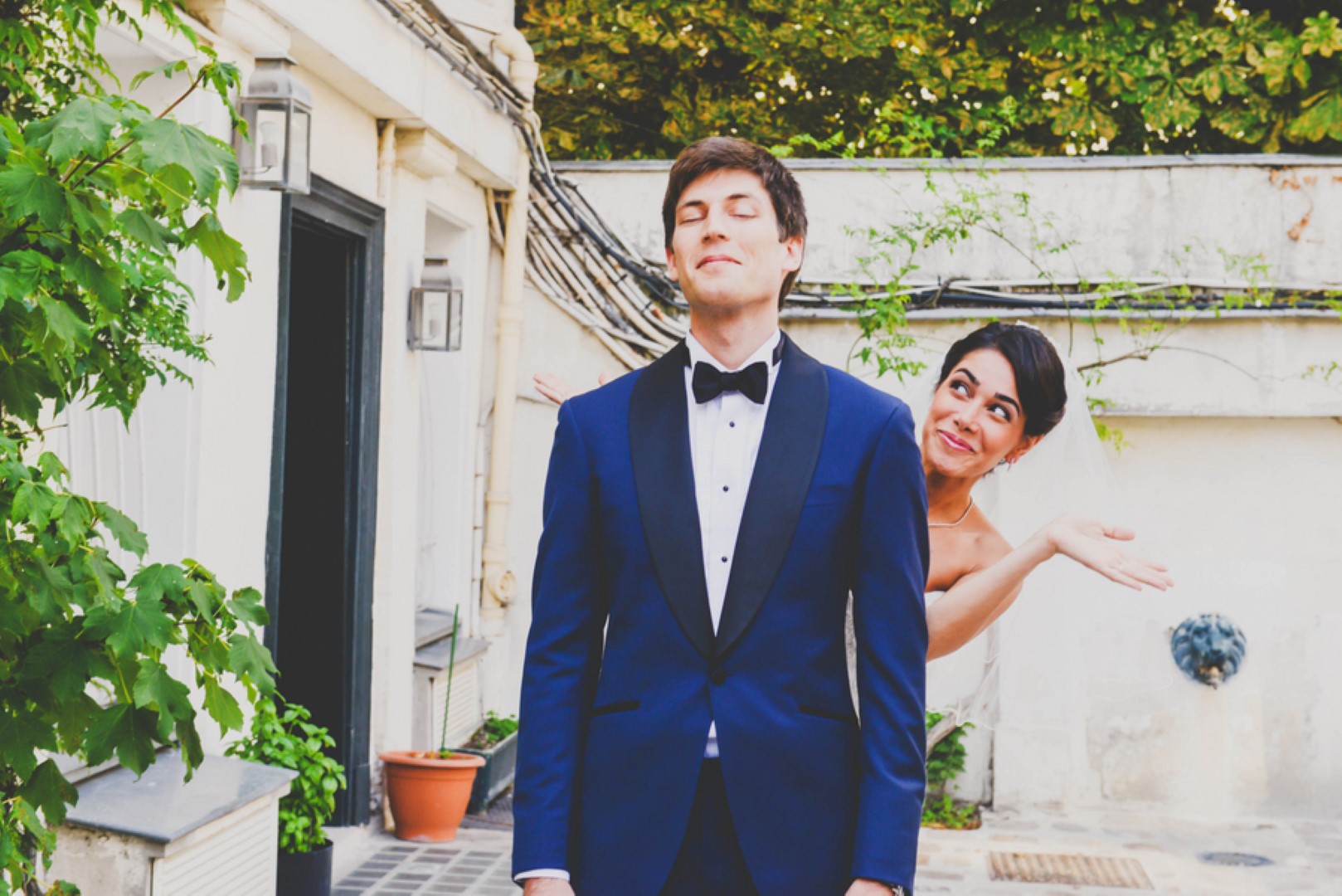 Groom waiting with his eyes closed while the bride peeks out from behind him with a playful shrug, in an ivy-covered Paris courtyard