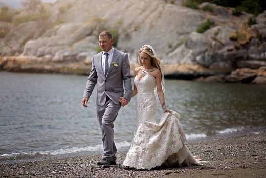 Bride and groom walking hand in hand along a rocky lakeshore, the groom in a grey suit
