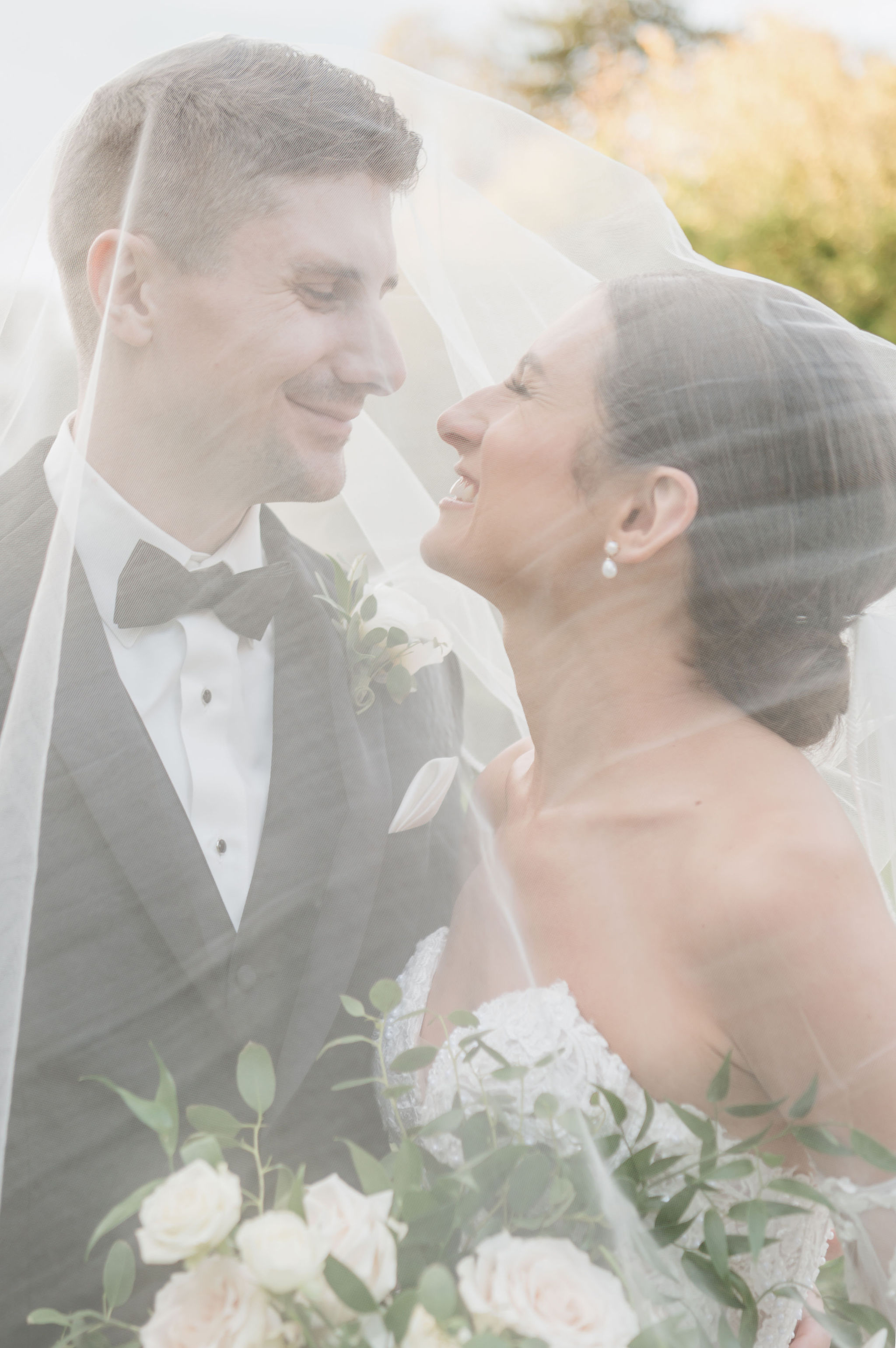 Bride's sheer veil draped over the couple as they smile and gaze at each other outdoors