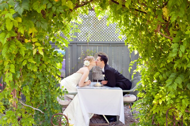 Bride and groom leaning across a small table to kiss under a leafy vine archway