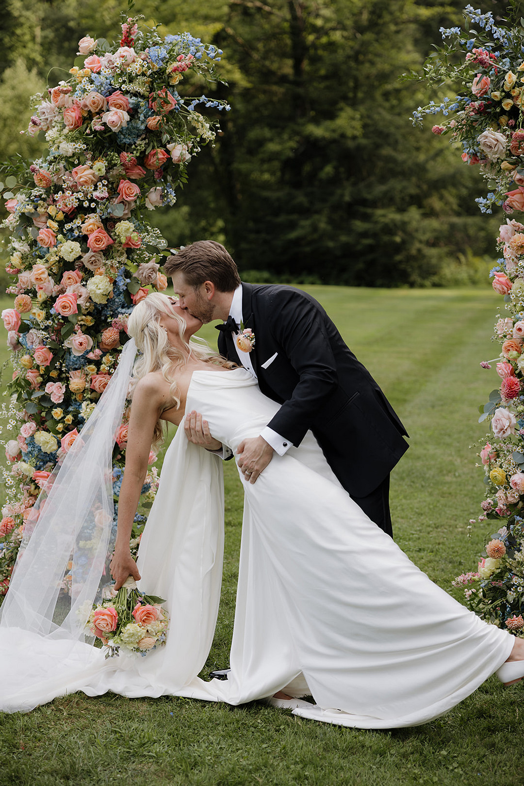 Groom dipping the bride backward into a kiss before a colorful floral ceremony arch