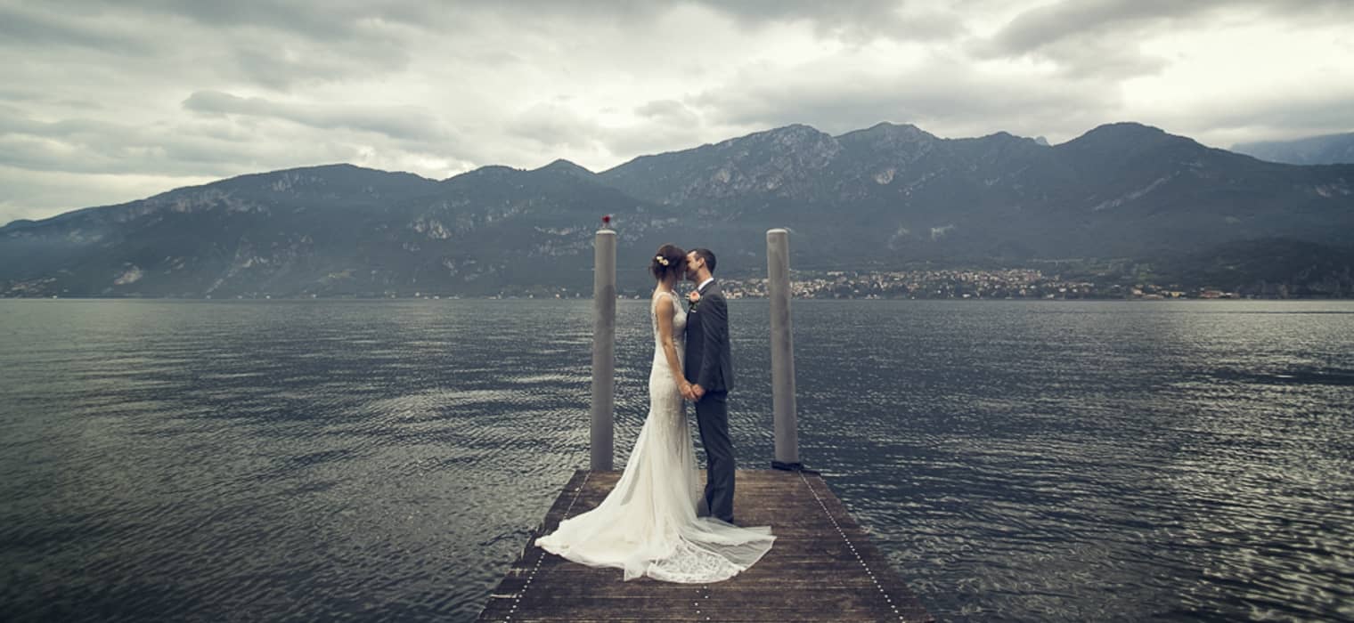 Bride and groom kissing on a wooden pier against a moody lake and mountain backdrop