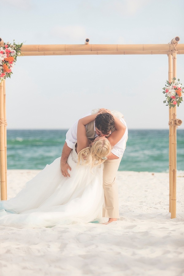 Groom dipping the bride backward into a kiss on a white-sand beach under a bamboo arch