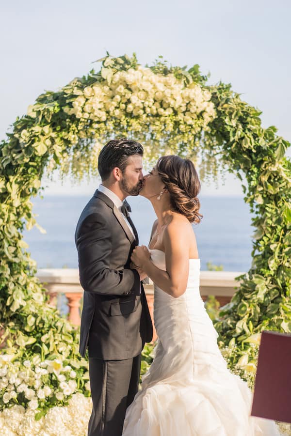 Couple kissing beneath a white floral arch with the ocean in the background