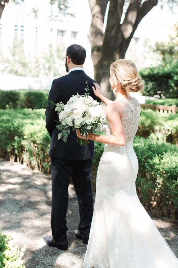 Bride in a lace gown reaching to tap the groom's shoulder from behind in a manicured boxwood garden, holding a white and greenery bouquet