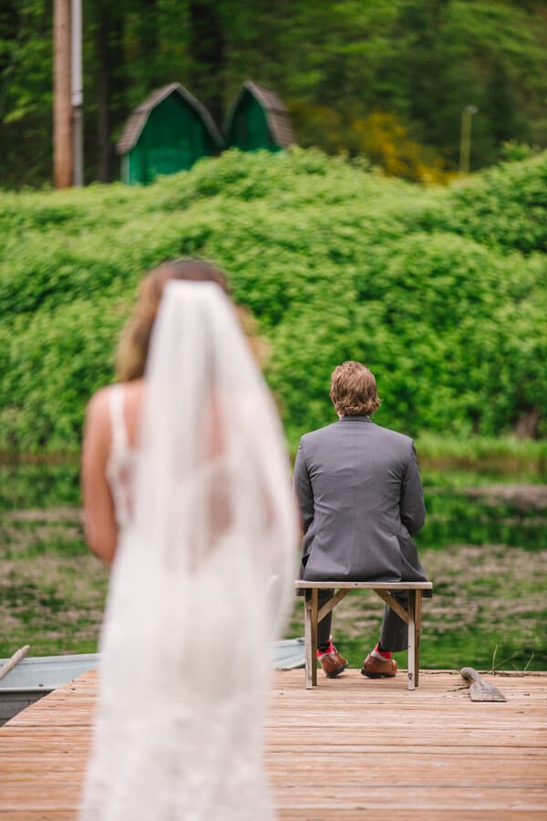 Groom seated on a wooden bench at the end of a pier facing a green pond, red socks showing, as the bride approaches seen over her veil